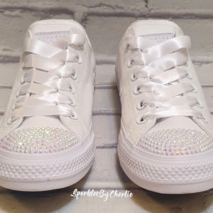 May include: A pair of white canvas sneakers with white satin laces and a row of iridescent rhinestones on the toe. The sneakers are tied with a bow.