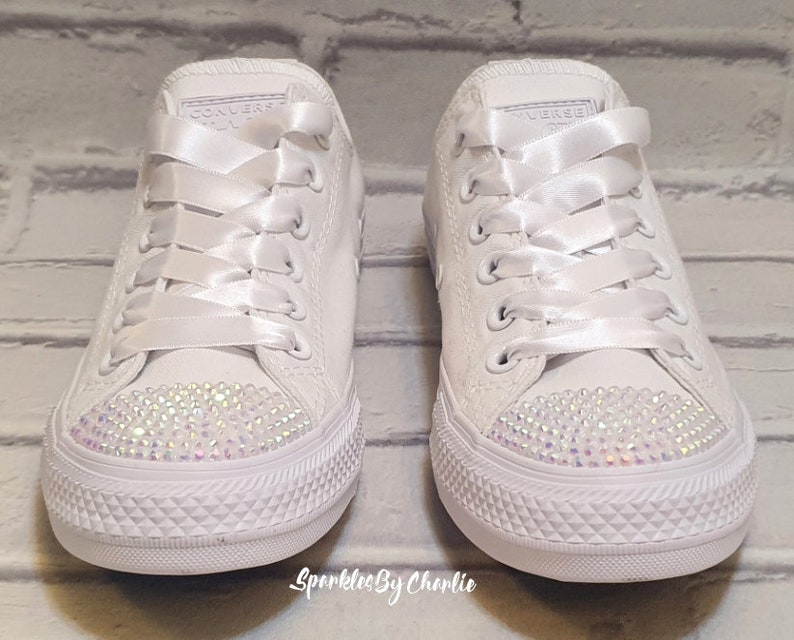 May include: A pair of white canvas sneakers with white satin laces and a row of iridescent rhinestones on the toe. The sneakers are tied with a bow.