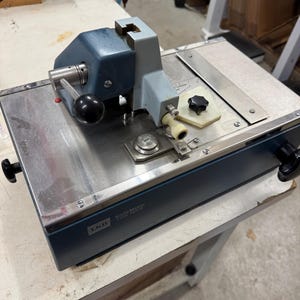 May include: A blue and silver metal machine with a handle and a knob. The machine has a label that reads "Kolle-Maker Type 7501R".
