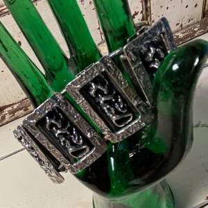 May include: A silver bracelet with a textured, geometric design. The bracelet is made up of multiple linked sections, each with a different pattern.
