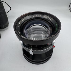 Op de afbeelding: Een zwarte cameralens met de tekst "Symmar 1:6.8/355" en andere markeringen. De lens heeft een groot, helder glaselement en een zilveren instelring. Aan de zijkant is een rode knop zichtbaar.