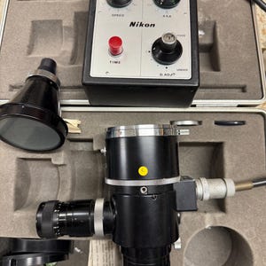 Könnte beinhalten: Ein schwarzes Nikon Kamera-Komponentenset, einschließlich eines Bedienfelds mit Drehreglern und Tasten mit der Aufschrift 'Speed', 'ASA' und 'Time'. Das Set enthält auch ein zylindrisches Objektiv und andere Komponenten, alles in einem grauen Koffer.