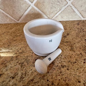 以下が含まれることがあります： 白のセラミック製乳鉢と乳棒のセット。乳鉢は注ぎ口が付いた丸いボウル型をしています。乳棒は円筒形で、丸い頭が付いています。どちらの部品にも小さな濃い色のロゴがあります。セットはまだらの茶色の表面にあります。