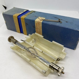 May include: A white plastic case with a clear glass bulb inside. The bulb has a metal base and a small metal wire attached to it. The case is open and the bulb is visible. The case is labeled "Manufactured by"