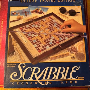 Pode incluir: Jogo de tabuleiro Scrabble Deluxe Travel Edition. A caixa azul apresenta o título do jogo em escrita branca. O tabuleiro está aberto, com uma mão colocando uma peça de letra. Óculos de sol e outros acessórios de jogo são visíveis.