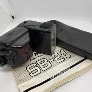 May include: A black Nikon SB-24 autofocus flash unit with a black carrying case and instruction manual. The flash unit has a red light on the front.