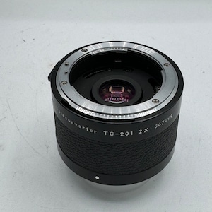 May include: A black and silver teleconverter lens, model TC-201 2X, with a textured black grip. The lens has a silver mounting ring and a lens element visible inside. The serial number 867499 is printed on the side.
