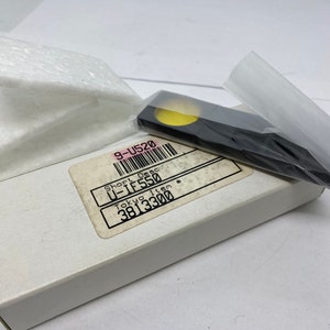 Puede incluir: Una caja blanca con una etiqueta en blanco y negro que dice "Short Desc U-IF550 Tokyo Item 3813300". La caja está sobre una superficie blanca. Un pequeño rectángulo negro con un círculo amarillo en el centro está sobre la caja.