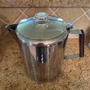 May include: A stainless steel coffee pot with a glass lid and a wooden handle. The pot has a spout for pouring and measurement markings on the side. The pot is sitting on a speckled countertop.