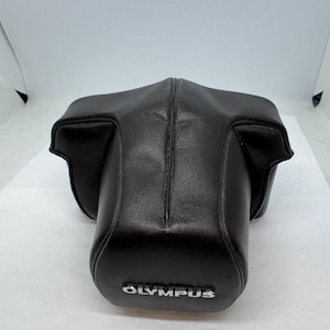 May include: Black leather camera case with the brand name "OLYMPUS" in silver lettering. The case is designed to fit a camera body and has a molded shape with a central seam. The case is made of a smooth, durable material.