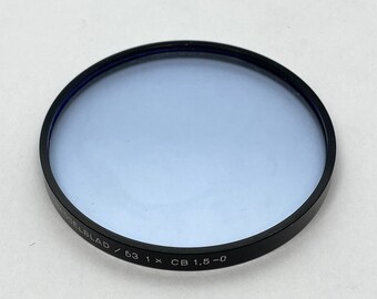 Könnte beinhalten: Ein schwarzer und silberner Filter für eine Hasselblad-Kamera. Der Filter hat einen Durchmesser von 63 mm und eine blaue Tönung. Der Text "HASSELBLAD / 63 1 x CB 1,5 - 0" ist auf dem Filter aufgedruckt.
