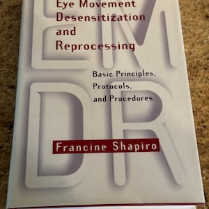 Könnte beinhalten: Ein Buch mit dem Titel "Eye Movement Desensitization and Reprocessing" von Francine Shapiro. Das Cover zeigt die Buchstaben "EMDR" in groß, weiß und schattiert. Der Titel ist in roter und schwarzer Schrift.