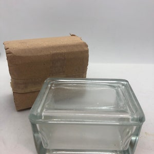 Puede incluir: Una caja de vidrio transparente con tapa. La caja es cuadrada y tiene un diseño simple. La caja está sobre una superficie blanca.