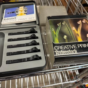 Könnte beinhalten: Ein Satz schwarzer Metallfilter in einem grauen Kunststoffgehäuse mit dem Text "Rodenstock CREATIVE PRINT SET". Eine Schachtel Rodenstock CREATIVE PRINT Filter mit einem Foto von grünen Pflanzen mit Tautropfen.