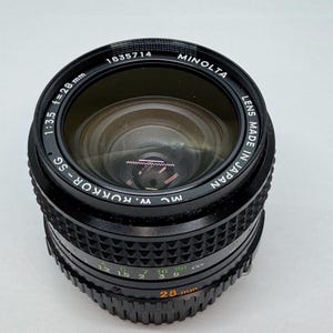 Puede incluir: Un objetivo de cámara Minolta negro con el texto "MINOLTA" y "LENS MADE IN JAPAN". El objetivo tiene una distancia focal de 28 mm y una apertura de f/3.5. El objetivo tiene marcas de medición.