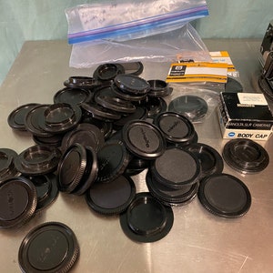 Puede incluir: Una colección de tapas de lentes y tapas de cuerpo de cámara Minolta negras. Las tapas están esparcidas sobre una superficie metálica, algunas aún en su embalaje original. Una bolsa de plástico es visible en el fondo.