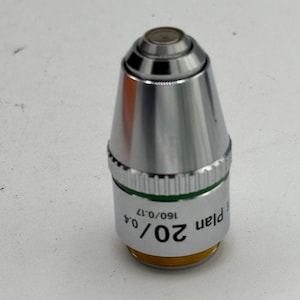 May include: A silver-colored microscope objective with a conical shape. The objective has a black top and a green band with white text that reads "20/0.4 160/0.17 Plan". The base is gold-colored.
