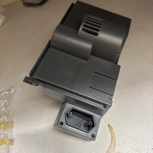 May include: A dark gray plastic component with a rectangular base and a curved, cylindrical section. The component has a series of ventilation slots and an electrical socket. The object is on a light-colored surface.