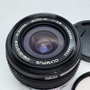 May include: Black Olympus OM-SYSTEM H.ZUIKO AUTO-W 1:2.8 f=24mm camera lens. The lens has a black body with white lettering and a silver aperture ring. The lens cap and filter are also visible.