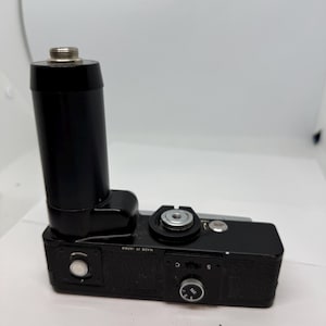 Può includere: Una macchina fotografica nera con un accessorio per obiettivo nero. La macchina fotografica ha un quadrante argentato con le lettere "S" e "C" e il testo "MADE IN JAPAN".