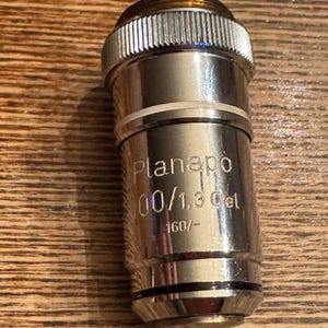 May include: A silver microscope objective lens with the text "Planapo 100/1.3 Oil 160/-". The lens has a ribbed top and a smooth, cylindrical body. The lens is designed for use in microscopy.