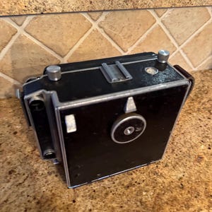 May include: A vintage black camera with silver accents. The camera has a rectangular body with a lens and various dials and knobs. The camera is sitting on a speckled brown surface.