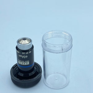 May include: A black and silver microscope objective lens with the text "ZEISS", "West Germany", "40/0.65", "97 00 - 066" printed on the side. The lens is in a clear plastic container.
