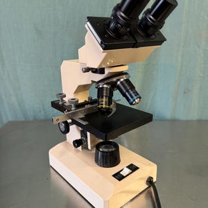 May include: A beige and black compound microscope with two eyepieces and a black stage. The microscope has a black power cord and a white switch on the base.