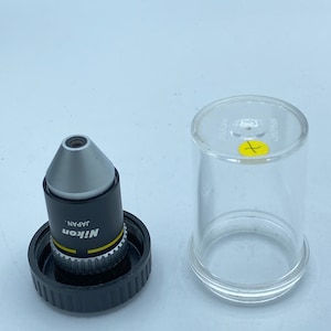 May include: A silver microscope objective lens with a black cap and a yellow label that reads "Nikon Japan". The lens is in a clear plastic storage tube.
