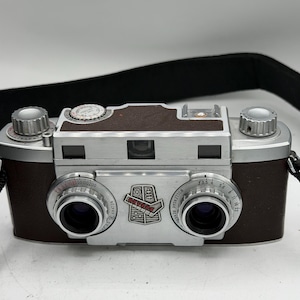May include: A vintage Revere stereo camera featuring a dual-lens system with silver metal housing and dark brown textured body panels. The front displays the Revere logo, two circular lenses with aperture and shutter speed markings, and a black strap.