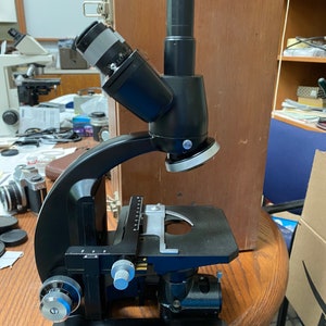 May include: A black microscope with a silver stage and a black and silver objective lens. The microscope is on a wooden table.