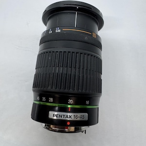 Pode incluir: Uma lente de câmera Pentax 16-45 preta. A lente tem um corpo canelado com marcações verdes e brancas. A lente tem uma tampa preta e vários indicadores de medição.