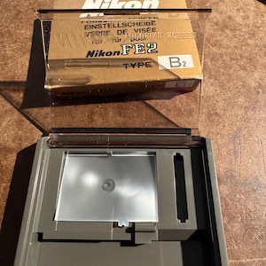 May include: A Nikon FE2 Type B2 focusing screen kit. The kit includes a clear plastic case with a gray base, a clear plastic cover, and a cardboard box with Nikon branding and text in multiple languages.