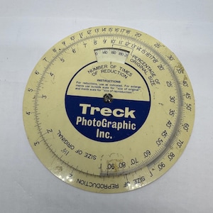 May include: A vintage Treck PhotoGraphic Inc. circular slide rule. The cream-colored tool features black and blue text, with scales for reduction and reproduction sizes. It is a tool for calculating percentages.