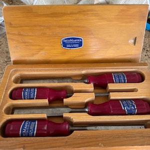 May include: A wooden box set containing four chisels. The chisels have dark red handles and silver blades. The box has a light wood finish and a blue and white label that reads "TECOMASTER PROFESSIONAL MADE IN SWEDEN EATONS OF CANADA."