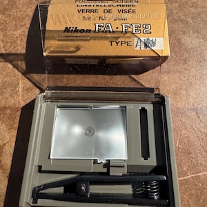May include: A Nikon FA FE2 Type A focus screen kit. The kit includes a clear plastic case, a brown cardboard box with the Nikon logo and text, and a gray plastic frame with a focus screen and black components.