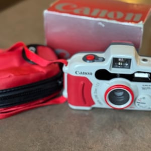 May include: A light gray and red Canon camera with a matching red carrying case and a red and white Canon branded box. The camera features a prominent red shutter button, a lens with a red ring, and the Canon logo printed on the front body.