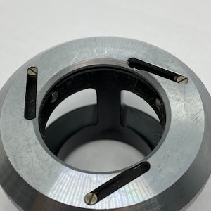 May include: A silver metal camera lens adapter with three slots for attaching filters. The adapter has a threaded ring on the bottom.