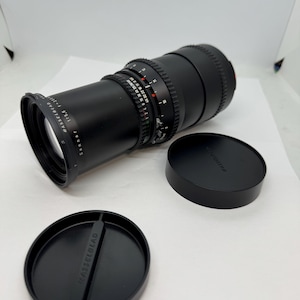 May include: A black Hasselblad Sonnar 1:5.6 f=250mm camera lens with two black lens caps. The lens has various measurement markings and a focus ring. The lens caps are round and feature the Hasselblad logo.