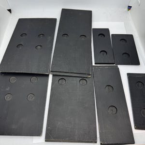 May include: A collection of black rectangular wooden dominoes with circular indentations. The dominoes vary in size and are arranged on a white surface. The set includes dominoes with two, three, and six indentations.