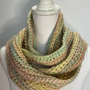 May include: A handmade, pastel-colored infinity scarf. The scarf is crocheted with a mix of soft colors, including light green, yellow, and peach. The circular design is displayed on a white mannequin bust, showcasing the texture and pattern of the yarn.