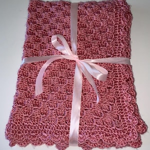 May include: A rectangular, dusty rose-colored crochet blanket featuring a repeating cluster stitch pattern. The edges are finished with a scalloped border. A light pink satin ribbon is tied in a bow around the center of the folded textile.