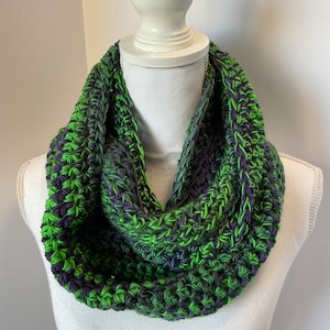 May include: A handmade infinity scarf in a vibrant color palette of green, purple, and gray. The chunky knit scarf is displayed on a white mannequin bust. The scarf is a cozy accessory for cold weather.
