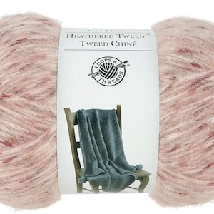 May include: A large ball of yarn in a soft pink hue with flecks of red and white. The label reads "Heathered Tweed Tweed Chine" with a logo of a yarn ball and needles. A teal blanket is draped over a wooden chair.