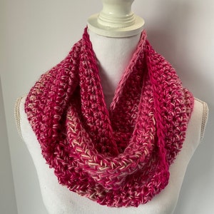 May include: A handmade, pink and white crocheted infinity scarf. The scarf is displayed on a white mannequin bust. The yarn has a textured appearance with varying shades of pink and white, creating a visually appealing accessory.