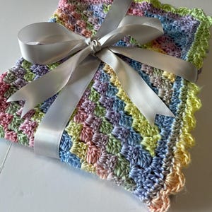 May include: A handmade, pastel-colored crochet blanket with a scalloped edge, tied with a silver satin ribbon. The blanket features a mix of pink, yellow, blue, and green yarn, creating a soft, textured appearance. Ideal for a baby.