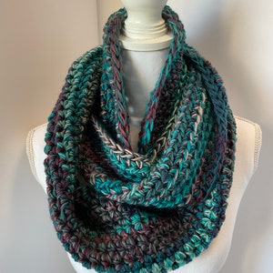 May include: A chunky, hand-knitted infinity scarf in shades of teal, burgundy, and cream. The thick yarn creates a textured, cozy look. The scarf is displayed on a white mannequin, showcasing its circular design and potential for warmth and style.