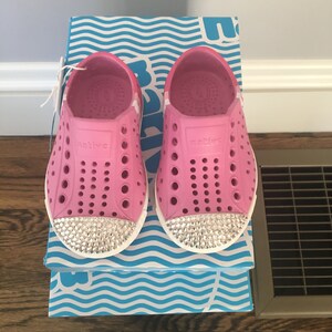 May include: A pair of pink children's shoes with white soles and rhinestones. The shoes are on a blue and white box with the word "Native" on them.