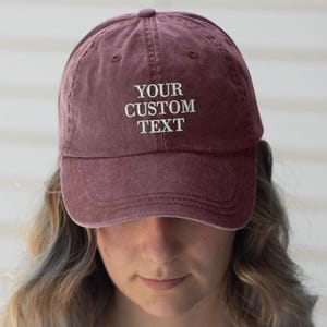 May include: A maroon baseball cap with the text "YOUR CUSTOM TEXT" embroidered in white. The cap has a curved peak and a slightly faded appearance, suggesting a vintage or worn look. The hat is a classic style, suitable for casual wear.