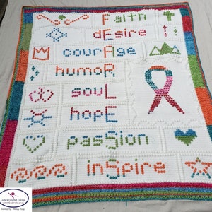 May include: A handmade white crochet blanket with colorful text and symbols. The blanket features words like "Faith," "Courage," and "Inspire." A pink, orange, and blue ribbon is also visible. The border is a mix of colors.